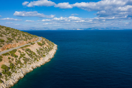 A winding road follows the edge of a rocky hillside covered with green shrubs, overlooking the expansive blue waters of the Peloponnese under a sky dotted with scattered clouds. The scene captures the raw beauty of coastal Greece and the contrast between rugged land and open sea.の写真素材