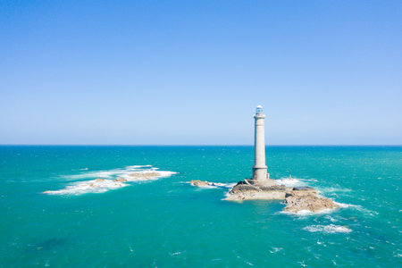A tall stone lighthouse stands alone on a rugged rocky islet, surrounded by vivid turquoise water and a clear blue sky at Cap de la Hague. The scene is open, bright, and evokes a sense of maritime isolation.の写真素材