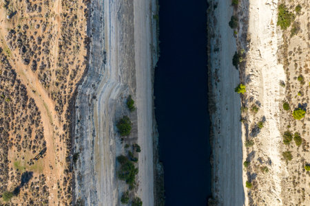 Direct overhead aerial of the Corinth Canal cutting through steep limestone walls, with sparse vegetation and dry, textured earth on both sides under strong sunlight.の写真素材