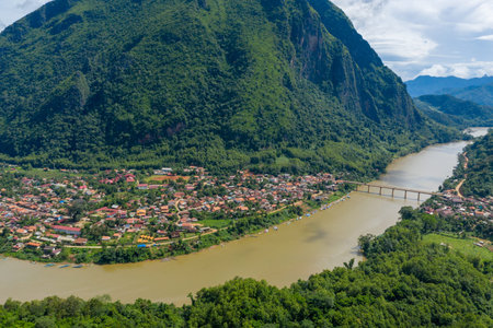 Wide aerial view of Nong Khiaw village in northern Laos, nestled between steep green limestone mountains and the Nam Ou river. The bridge, riverside houses, and dense tropical forest create a vibrant landscape under a bright summer sky.の写真素材
