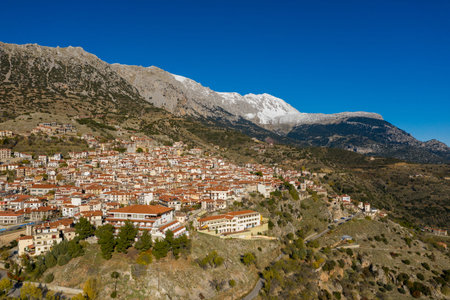 Panoramic aerial view of Arachova, a Greek mountain village with red-tiled roofs nestled on a rugged hillside, framed by dramatic snow-capped peaks and a deep blue sky.の写真素材