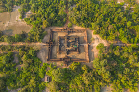Drone perspective reveals the full layout of Banteay Samre temple surrounded by dense forest and rural clearings in the Angkor region of Cambodia. The ancient sandstone structure stands out against the lush greenery under warm daylight.の写真素材