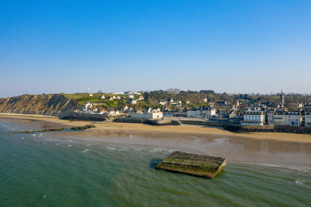 Drone perspective of Arromanches-les-Bains reveals sandy beach, historic concrete structure in shallow water, dramatic cliffs, and charming village architecture beneath a vibrant blue sky.の写真素材