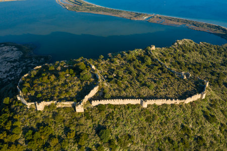Aerial image of ancient stone castle walls enclosing a green, rugged hilltop with sweeping views of a blue coastal lagoon near Navarino, Greece, in bright sunlight.の写真素材