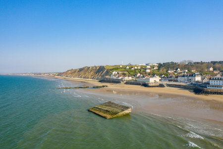 Aerial view of Arromanches-les-Bains with sandy beach, calm sea, and dramatic cliffs under a clear blue sky. The historic concrete structure in the water adds visual interest to the tranquil coastal scene.の写真素材