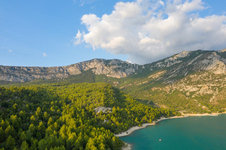 Dense pine woods meet the turquoise shoreline of Lac de Sainte-Croix, with rugged limestone mountains rising in the background beneath a sky dotted with sunlit clouds.の写真素材