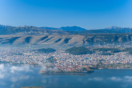 A wide panoramic scene captures the city of Ioannina nestled beside Lake Ioannina, with the dramatic ridges of the Pindus mountains rising in the background. Low clouds drift above the calm water, highlighting the natural textures and urban landscape of Epirus, Greece.の写真素材