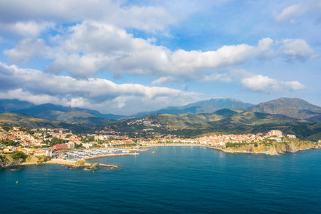 Aerial panorama of Banyuls-sur-Mer showing a vibrant coastal town, sheltered marina, and dramatic mountains under a sky dotted with clouds and soft sunlight.の写真素材