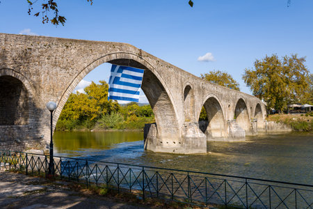 Historic arched stone bridge spans a clear river in Arta, Greece. The scene features textured masonry, autumn trees, and blue sky in gentle daylight.の写真素材
