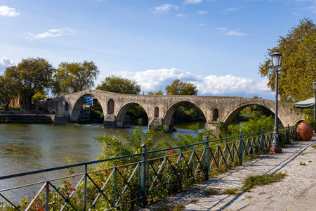 Historic multi-arched stone bridge spans a tranquil river in Arta, Greece. The foreground features an iron fence, leafy plants, and a cobblestone walkway under clear daylight, creating a peaceful riverside scene.の写真素材