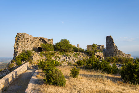 Weathered stone walls of Chateau de Opoul Perillos stand atop a rugged hill, surrounded by wild grasses and shrubs in warm afternoon light under a clear blue sky.の写真素材