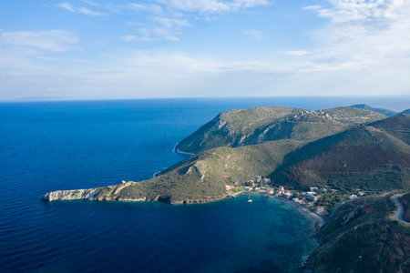 Aerial photograph captures a tranquil bay with a small coastal village nestled at the base of rugged green hills on a dramatic peninsula in Magne, Greece. The deep blue Mediterranean Sea surrounds the land, while soft daylight highlights the natural contours and textures.の写真素材