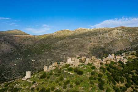 Aerial view of the historic stone village of Vathia perched on a lush hillside, surrounded by rugged mountains and clear blue sky. The old towers and clustered buildings create a striking contrast with the wild Mediterranean landscape.の写真素材