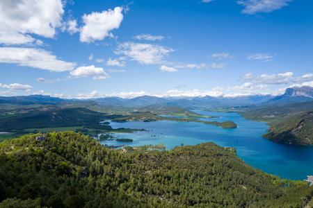 Sweeping view of Mediano Lake in Aragon, Spain, with vivid turquoise water winding between lush green hills and distant snow-capped Pyrenees under a bright blue sky dotted with clouds. Sunlight enhances the vibrant colors and natural textures.の写真素材