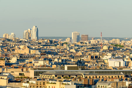 Expansive view over Paris rooftops with a mix of historic low-rise buildings and distant modern skyscrapers, bathed in warm evening sunlight under a clear sky.の写真素材