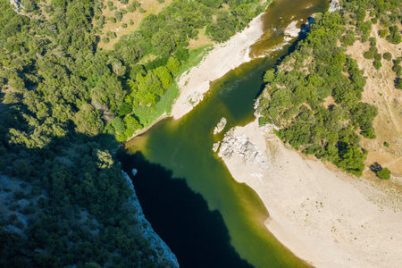 Aerial view of a winding river bordered by rocky banks and dense green woodland in the Gorges de lArdeche. Sunlight casts sharp contrasts on the water and surrounding terrain.の写真素材