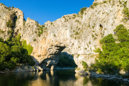 Towering natural stone arch spans a serene river, surrounded by rugged cliffs and vibrant green foliage at Pont dArc in Ardeche. Clear blue sky and golden sunlight highlight the dramatic landscape.の写真素材