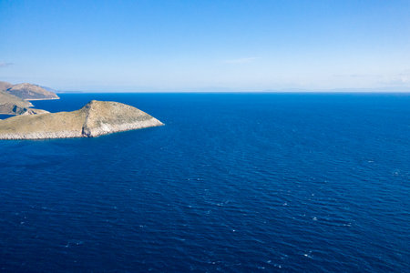 Aerial view of a sunlit, rocky promontory jutting into the vast, deep blue Mediterranean at Cap Tenare, Greece. The scene conveys openness and tranquility, with rugged terrain and expansive water under a clear sky.の写真素材