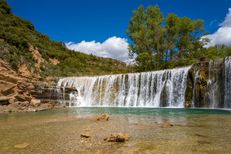 Crystal-clear waterfall tumbles over a wide stone ledge into a shallow turquoise pool, bordered by rugged rocks and vibrant green trees under a bright blue sky in Bierge, Spain. Sunlight enhances the sparkling water and natural landscape.の写真素材