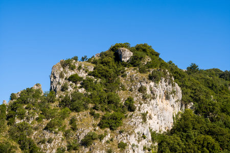 Steep limestone outcrop covered in lush green shrubs and trees rises against a vivid clear blue sky in the Gorges de lArdeche. Sunlight highlights rugged rock textures and natural vegetation.の写真素材