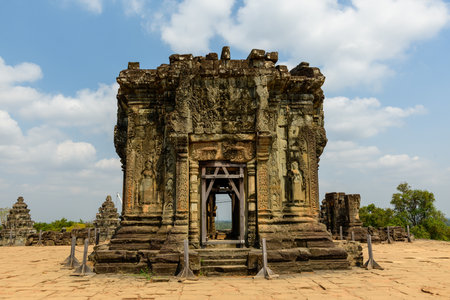 The main stone shrine of Phnom Bakheng temple stands on a raised terrace near Siem Reap, Cambodia, featuring intricate carvings and weathered bas-reliefs under a bright sky. The ancient structure is surrounded by open paving and distant greenery, highlighting its historic and monumental character.の写真素材