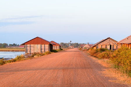 Warm sunlight illuminates rustic wooden salt storage sheds with red metal roofs along a wide red dirt road near Kampot, Cambodia. The tranquil rural scene features open salt ponds, scattered vegetation, and distant palm trees under a pale sky.の写真素材