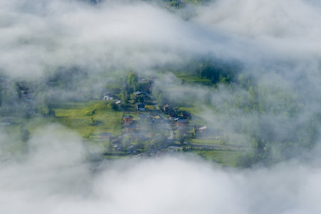 A cluster of alpine houses and green fields appears through drifting low clouds, with soft sunlight highlighting the lush landscape near Chamonix. The scene feels peaceful and secluded, wrapped in early morning fog.の写真素材