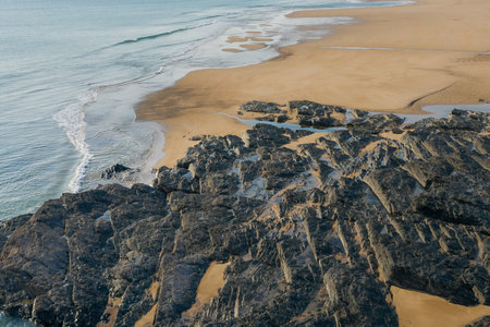 Jagged dark rocks form intricate tidal pools along a wide sandy shoreline at Cap de Carteret, Normandy. Soft morning light highlights the textures of the rocks and the gentle waves meeting the beach.の写真素材