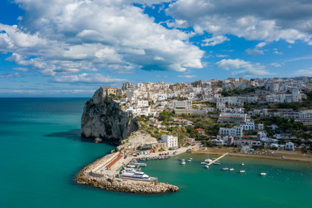 Dramatic aerial view of Peschici with white buildings clustered on a steep cliff above a vibrant turquoise harbor, set against a dynamic sky with scattered clouds.の写真素材
