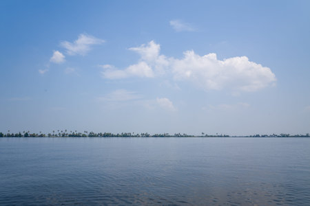 Wide tranquil backwater stretches beneath a vast blue sky in Alappuzha, India, with a distant line...
