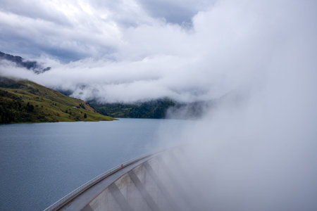 Low clouds and mist sweep across the curved concrete structure of Roselend Dam and the calm blue waters of the alpine reservoir, with green mountain slopes fading into the fog. The scene features diffused light and a tranquil, atmospheric mood in the French Alps.の写真素材