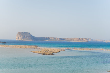 A tranquil sandbar stretches into shallow turquoise water with a distinctive flat-topped island in the distance near Balos, Crete. The scene is sunlit and serene, with soft blue tones and a clear sky.の写真素材
