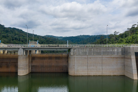 A large concrete dam structure spans a calm reservoir, set against a background of dense green...