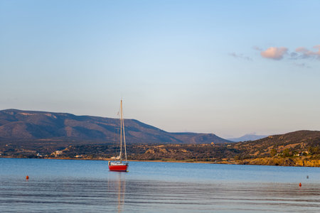 A single red sailboat floats in calm blue water, surrounded by rugged hills and distant mountains...