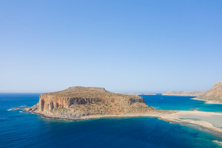 A rugged rocky peninsula rises from vivid azure waters at Balos Beach in Crete, Greece. The landscape is sunlit and expansive, with clear skies and contrasting earth tones.の写真素材