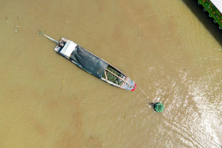 Overhead image captures a single long wooden boat loaded with green produce, covered by a tarpaulin,...