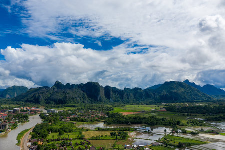 Sweeping aerial view of Vang Vieng, Laos, with dramatic limestone cliffs rising behind a patchwork...