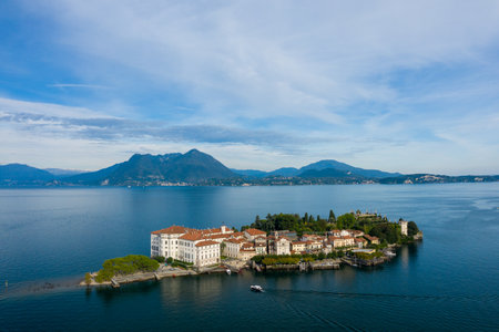 Aerial perspective of Isola Bella surrounded by the deep blue waters of Lake Maggiore, with a grand palace and manicured gardens in the foreground. Alpine mountains and lakeside towns form a scenic backdrop beneath a sky streaked with soft clouds in northern Italy.の写真素材