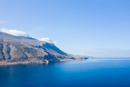 Bright sunlight illuminates rugged, rocky hills meeting calm, deep blue sea at Pointe de Balos, Crete. The expansive sky and tranquil water create a sense of peaceful openness.の写真素材