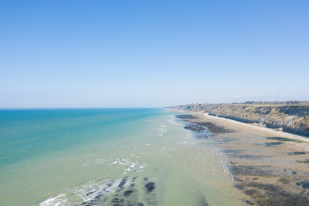 Wide aerial view of a sandy shoreline with dramatic cliffs and turquoise sea near Port en Bessin, Normandy. Clear blue sky and tranquil water create a serene coastal atmosphere.の写真素材