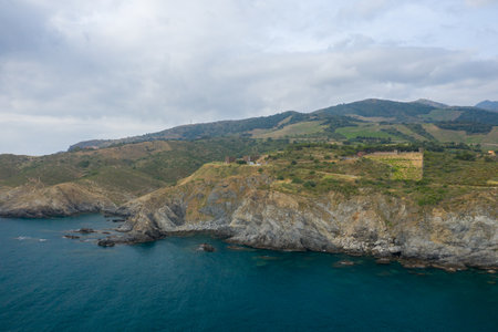 Aerial view of a rocky coastal headland rising above deep blue Mediterranean waters, with green hills and terraced slopes under a cloudy sky near Banyuls-sur-Mer.の写真素材