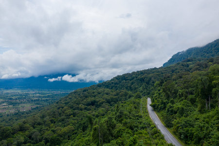 A narrow paved road curves along lush green hillsides covered in dense tropical forest, with...