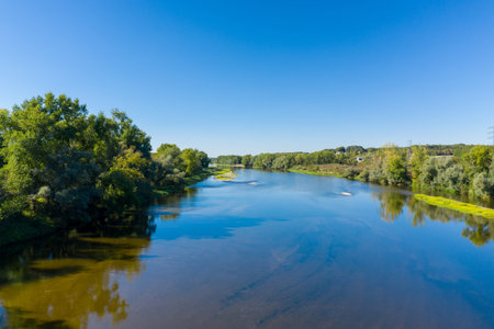 Wide river bordered by dense green trees and sandy patches flows through open countryside beneath a...