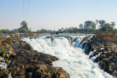 Energetic whitewater plunges over rugged stone ledges at Tat Somphamit Waterfalls, also known as Li Phi Falls, in Champasak Province, southern Laos. The scene features textured rocks, swirling rapids, and sparse greenery beneath a clear morning sky.の写真素材