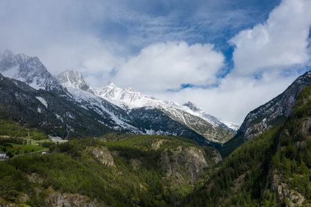 Snow-covered mountain peaks rise above dense green forests and rocky hillsides in Val dAoste, Italy....