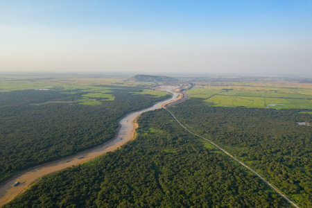 Aerial view of a muddy river curving through dense green forest and patchwork rice fields near Tonle Sap in Cambodia. The expansive landscape features distant hills, open countryside, and a clear blue sky, evoking a sense of tranquility and natural beauty.の写真素材