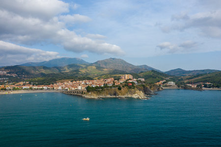 Aerial view of Banyuls-sur-Mer perched on rocky cliffs above the blue Mediterranean Sea, with rolling green hills and dramatic mountains in the background under a partly cloudy sky.の写真素材