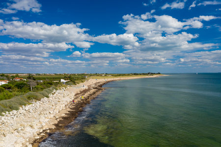 A sweeping view of a sandy beach edged by a rocky shoreline and green vegetation on Ile de Re, Charente-Maritime. Puffy white clouds drift across a vivid blue sky above calm Atlantic waters, creating a spacious and tranquil coastal atmosphere.の写真素材