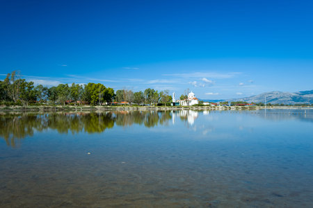 A white church and a line of green trees are mirrored in the calm water of Tourlida lagoon in western Greece. The scene features clear reflections, a vivid blue sky, and distant mountains, creating a peaceful wetland atmosphere.の写真素材