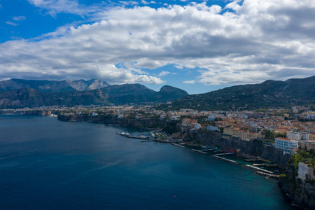 Sweeping clouds drift above the dramatic cliffs and marina of Sorrento, Italy, with the towns pastel buildings perched along the rugged shoreline. The scene captures deep blue Mediterranean water, mountainous backdrop, and a tranquil coastal atmosphere.の写真素材